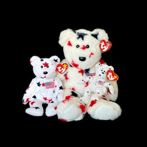Puede incluir: Tres osos de peluche blancos con patrones de estrellas rojas, blancas y azules. El oso más grande tiene una etiqueta Ty roja, blanca y azul en su oreja. Los dos osos más pequeños tienen una etiqueta Ty roja, blanca y azul en su oreja izquierda.
