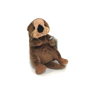 May include: A brown plush sea otter soft toy with a black nose and white whiskers. The otter is sitting up with its arms crossed and has a blue tag attached to its wrist.