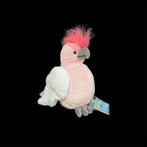 May include: Pink and white plush cockatoo bird with a grey beak and a pink crest. The cockatoo is sitting on a white surface with a blue and yellow tag that says "Webkinz"