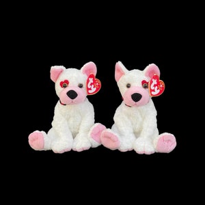 Peut inclure: Deux chiens en peluche blancs avec des nez et des pattes roses. Chaque chien a une étiquette rouge en forme de cœur avec le mot "TY" dessus.