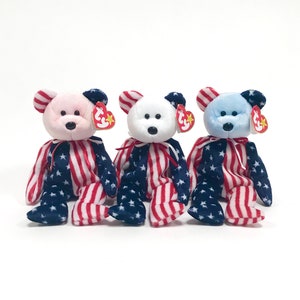 May include: Three plush teddy bears wearing American flag patterned outfits. The bears are pink, white, and blue.