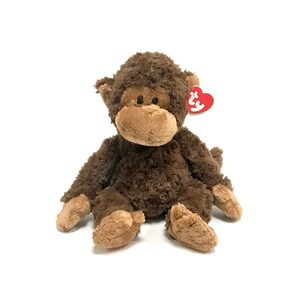 以下が含まれることがあります： 茶色の毛皮、ベージュの顔、赤いハート型のタグが付いたぬいぐるみのおもちゃ。ぬいぐるみは腕と脚を広げて座っています。猿はフレンドリーな表情をしています。