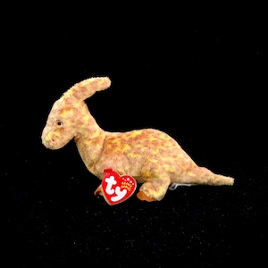 Pode incluir: Um brinquedo de dinossauro de pelúcia amarelo e marrom com uma etiqueta vermelha Ty Beanie Babies. O dinossauro tem um pescoço longo e uma cabeça pequena com um olho grande.