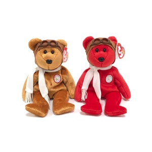 May include: Two plush beanie baby bears wearing aviator hats and scarves. The bear on the left is brown and the bear on the right is red.