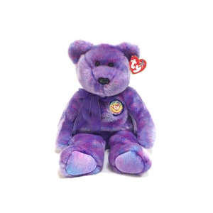 Può includere: Un orsacchiotto viola con un fiocco e un'etichetta a forma di cuore. L'orso ha un naso nero e un piccolo emblema circolare sul petto. La pelliccia è un mix di tonalità viola, con sfumature più chiare sui piedi.