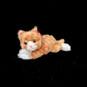 Op de afbeelding: Een pluche oranje tijgerkat speelgoed met witte poten en een roze neus. De kat heeft een bruin lint om de nek en een rood label aan het oor. Het speelgoed ligt op een zwarte ondergrond.