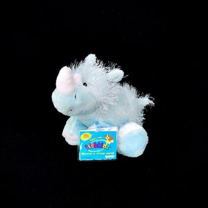 May include: A blue Webkinz plush rhino with a pink horn and a white belly. The rhino is sitting on a white surface with a Webkinz tag attached to its leg. The tag is blue, yellow, red, and green and says "Webkinz discover a virtual world! Ganz."