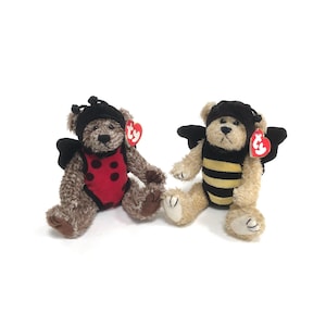 May include: Two plush teddy bears dressed as insects. One bear is dressed as a ladybug with red and black spots. The other bear is dressed as a bee with black and yellow stripes. Both bears have Ty tags attached to their costumes.