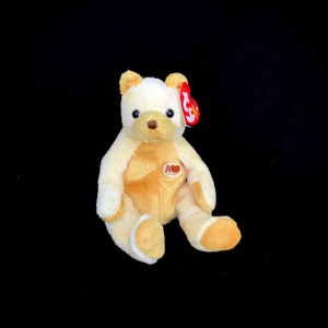 Puede incluir: Un oso de peluche crema y marrón con una etiqueta roja en la oreja. El oso tiene una nariz marrón y ojos negros. El peluche está sentado sobre un fondo negro. El oso tiene detalles marrones en el pecho, las patas y las orejas.