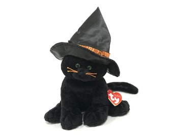 Puede incluir: Un juguete de peluche de gato negro con un sombrero de bruja negro con una banda a cuadros naranja y marrón. El gato tiene ojos verdes y bigotes naranjas. Una etiqueta roja en forma de corazón está adjunta.