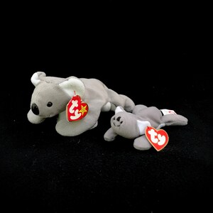 May include: Two grey plush koala beanie babies with red Ty tags. The larger koala is lying on its side with its head facing the left. The smaller koala is lying on its belly with its head facing the right.
