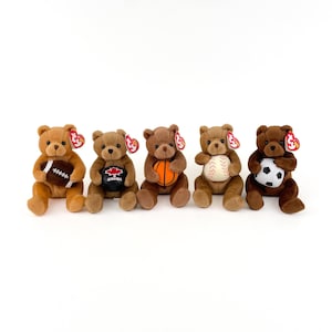 May include: Five brown teddy bears with red and white beanie baby tags. Each bear is holding a different sports ball: a football, a baseball, a basketball, a soccer ball, and a baseball.