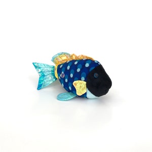Puede incluir: Un juguete de peluche con forma de pez, con un cuerpo azul cubierto de lunares blancos. El pez tiene una cabeza negra, una cola turquesa y aletas amarillas y doradas. El juguete está diseñado para parecerse a un pez tropical.