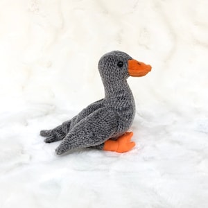 Puede incluir: Un ganso de peluche gris con pico y patas naranjas está sentado sobre una superficie blanca texturizada. El animal de peluche tiene ojos negros y un cuerpo suave y texturizado. Este ganso hecho a mano es un objeto decorativo encantador.