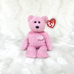 May include: Pink plush teddy bear with a white swan embroidered on its chest. The bear has a pink ribbon around its neck and a Ty Beanie Babies tag attached to its ear. The text "It's a girl" is embroidered on the bear's chest.