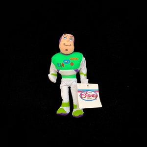 May include: A Buzz Lightyear plush doll from the Disney Store. The doll is wearing a green, white, and purple spacesuit with the words "LIGHT YEAR" on the chest. The doll has a tag attached with the Disney Store logo.