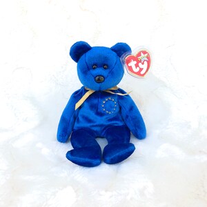 May include: A blue plush bear with a gold ribbon around its neck and a gold star design on its chest. The bear has a Ty heart-shaped tag attached. The bear is sitting on a white, textured surface.
