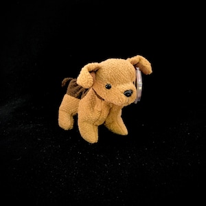 May include: A brown and tan plush toy dog with a brown patch on its back. The dog has a white tag on its ear.