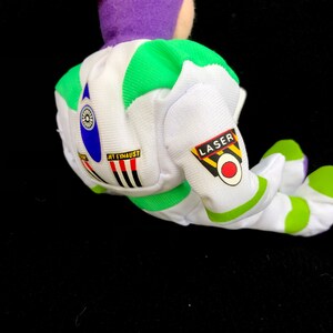 May include: A plush toy of a space ranger, featuring a white and green spacesuit with the words "JET EXHAUST" and "LASER". The toy has a purple head and green accents. The toy is lying on a black surface.