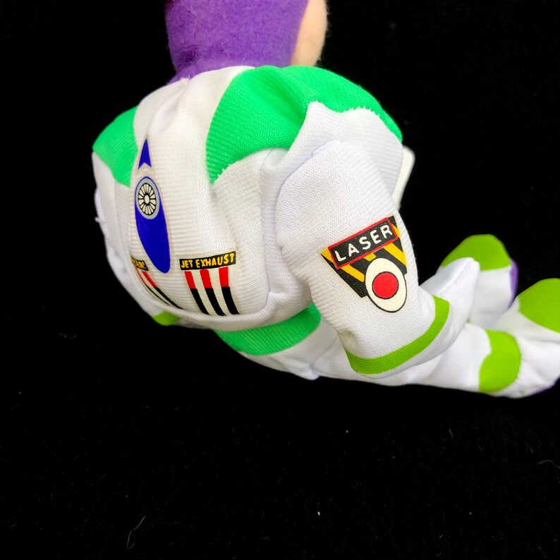 May include: A plush toy of a space ranger, featuring a white and green spacesuit with the words "JET EXHAUST" and "LASER". The toy has a purple head and green accents. The toy is lying on a black surface.