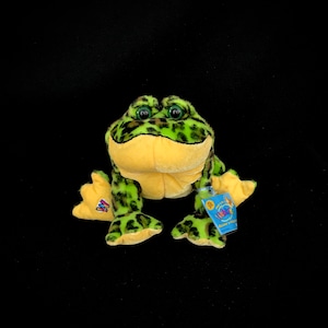 May include: Green and yellow frog plush toy with black spots. The toy has a Webkinz tag attached to its leg. The tag is blue and yellow with the words "Webkinz" and "Become a Member" printed on it.