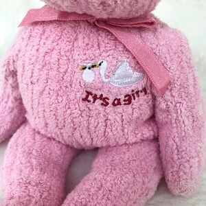 May include: Pink plush teddy bear with a white swan and the text "It's a girl" embroidered on the chest. The bear has a pink ribbon tied around its neck.