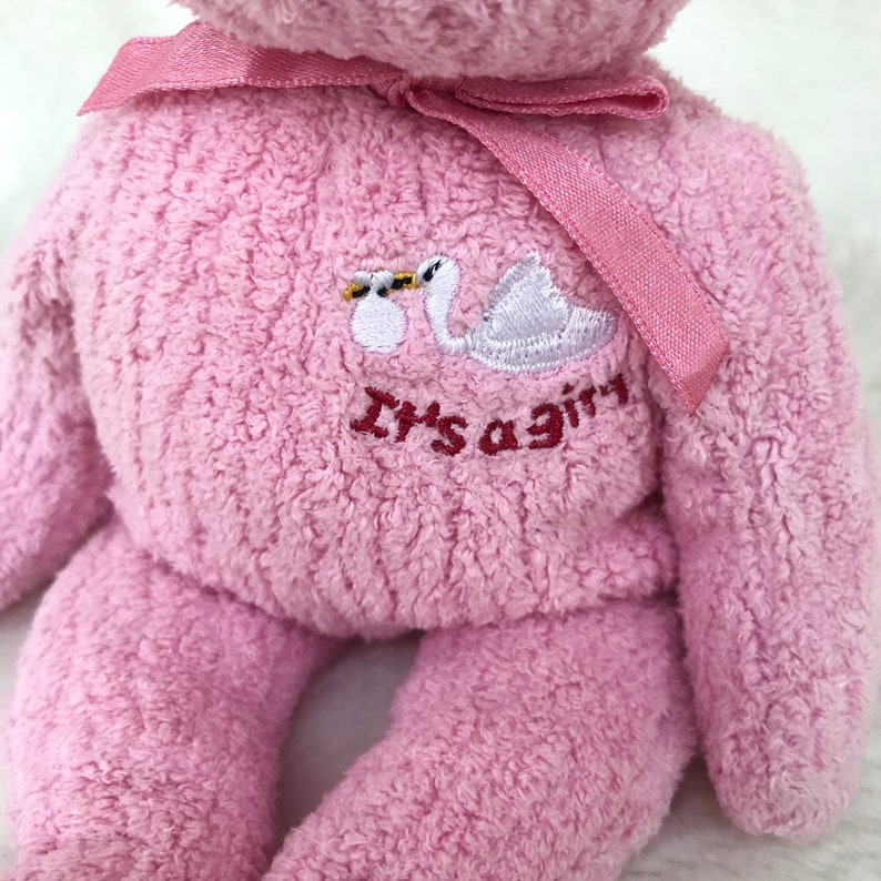 May include: Pink plush teddy bear with a white swan and the text "It's a girl" embroidered on the chest. The bear has a pink ribbon tied around its neck.