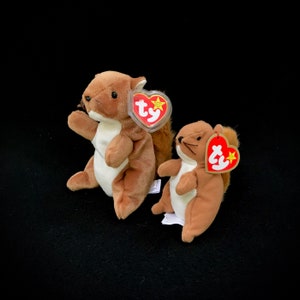 May include: Two brown and white squirrel beanie babies with red and white Ty tags. The squirrels are sitting on a black surface.