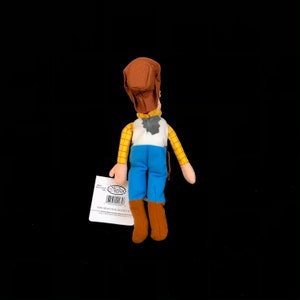 May include: A plush Woody doll from the Disney Pixar Toy Story franchise. The doll is wearing a brown cowboy hat, a yellow plaid shirt, a white vest, blue pants, and brown boots. A tag is attached to the doll.