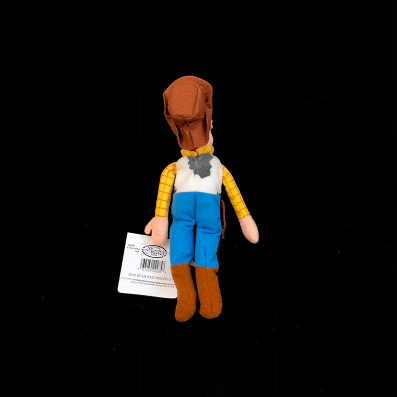 May include: A plush Woody doll from the Disney Pixar Toy Story franchise. The doll is wearing a brown cowboy hat, a yellow plaid shirt, a white vest, blue pants, and brown boots. A tag is attached to the doll.