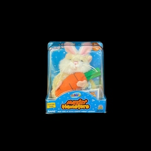 May include: A white plush hamster holding a carrot, wearing a pink bunny costume. The hamster is in a clear plastic package with a blue and white background. The package says "Webkinz Mazin' Hamsters" and "First Edition".