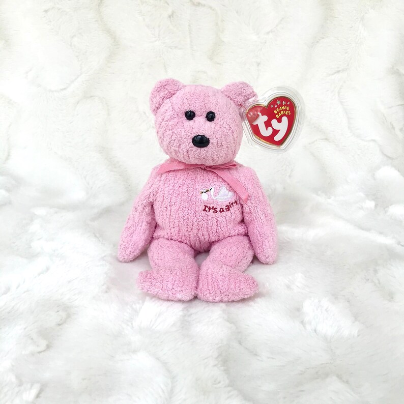 May include: Pink plush teddy bear with a white swan embroidered on its chest. The bear has a pink ribbon around its neck and a Ty Beanie Babies tag attached to its ear. The text "It's a girl" is embroidered on the bear's chest.