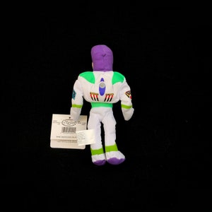May include: A plush Buzz Lightyear mini bean bag toy, viewed from the back, with a white and green spacesuit, purple hood, and tag. The tag reads "Mini Bean Bag Buzz". The toy is on a black background.