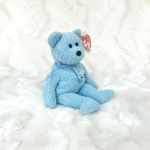 May include: A light blue plush teddy bear with a white ribbon and a Ty tag. The bear is sitting on a white fluffy surface.