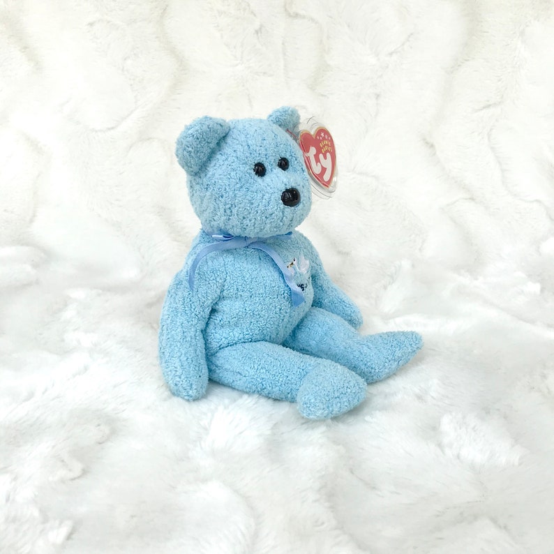 May include: A light blue plush teddy bear with a white ribbon and a Ty tag. The bear is sitting on a white fluffy surface.
