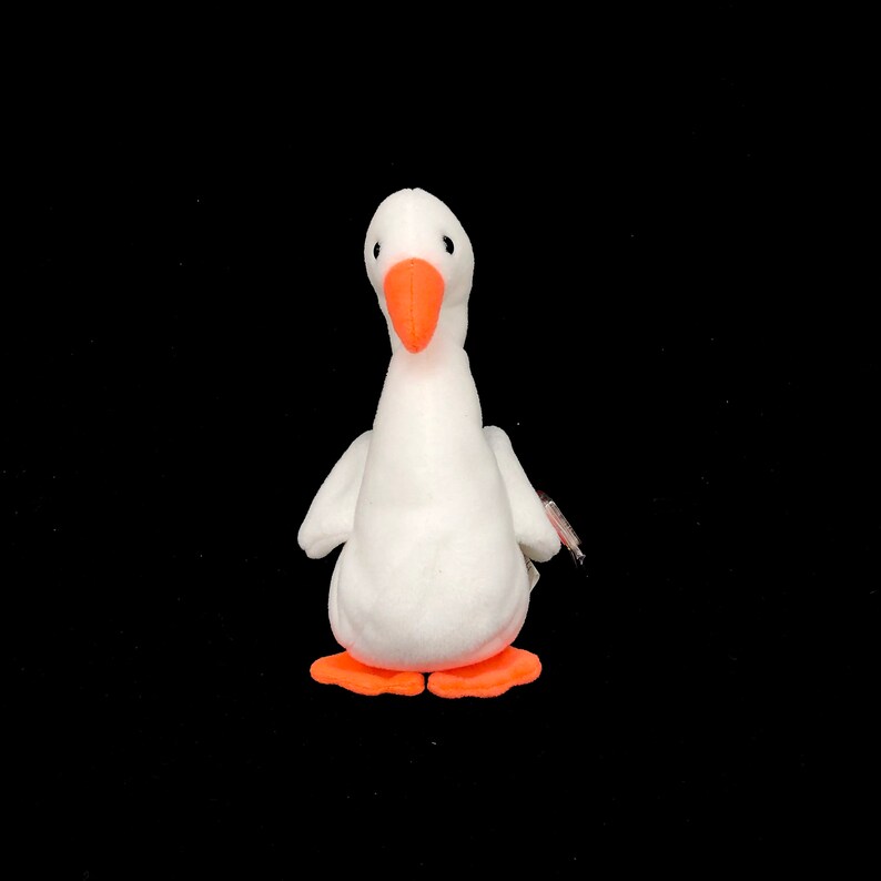 Op de afbeelding: Een witte pluche gans speelgoed met een oranje snavel en poten. Het knuffeldier heeft kleine zwarte ogen en een label aan de zijkant. De gans staat rechtop tegen een zwarte achtergrond.