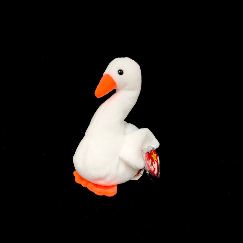 Op de afbeelding: Een witte pluche gans speelgoed met een oranje snavel en poten. De gans heeft een rood en geel label aan zijn vleugel. Het speelgoed staat tegen een zwarte achtergrond.