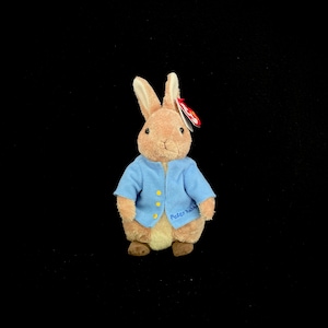 Puede incluir: Un juguete de peluche de Peter Rabbit con una chaqueta azul con botones amarillos. El juguete tiene una etiqueta Ty roja unida a su oreja. El juguete está de pie sobre un fondo negro.