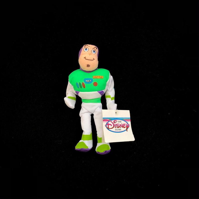 May include: A Buzz Lightyear plush doll from the Disney Store. The doll is wearing a green, white, and purple spacesuit with the words "LIGHT YEAR" on the chest. The doll has a tag attached with the Disney Store logo.