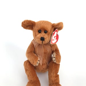 Puede incluir: Un oso de peluche marrón con nariz y ojos negros, y una cinta marrón alrededor del cuello. El oso tiene una etiqueta roja sujeta a la oreja. El peluche está sobre un fondo blanco.