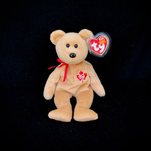 Könnte beinhalten: Ein brauner und beiger Beanie Baby Bär mit einem roten Band um den Hals. Der Bär hat ein rotes Ahornblatt auf der Brust und ein Ty-Etikett am Ohr. Das Etikett lautet "10th Anniversary Beanie Babies".