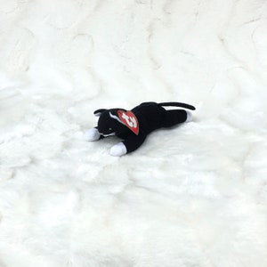 Op de afbeelding: Een zwart pluche kattenknuffel met witte poten en een witgepunte staart. De kat heeft groene ogen en een rood hartvormig label. Het speelgoed ligt op een wit, getextureerd oppervlak.