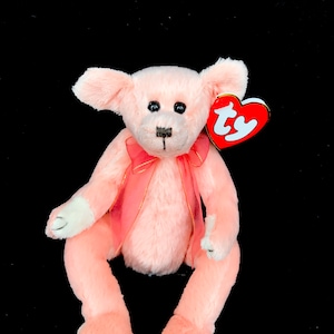 Puede incluir: Oso de peluche rosa con una cinta rosa alrededor del cuello y una etiqueta Ty roja unida a su oreja. El oso tiene una nariz blanca y ojos negros.