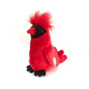 Puede incluir: Un juguete de peluche de un pájaro cardenal rojo, con detalles en negro. El juguete tiene una cresta roja prominente, una cara negra y un pico rojo. El pájaro está sentado, con una etiqueta adjunta en el lateral.