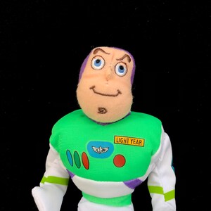 May include: A plush Buzz Lightyear toy from the Toy Story franchise. The toy features a green and white spacesuit with a "Light Year" patch, purple accents, and a smiling face with blue eyes. The toy is set against a black background.