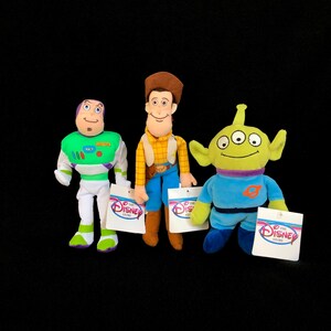 May include: Three plush Toy Story character dolls: Buzz Lightyear, Woody, and an Alien. Buzz is in a green and white spacesuit. Woody wears a cowboy outfit. The Alien is green with three eyes. Each doll has a Disney Store tag.