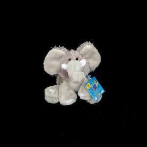 Puede incluir: Un peluche de elefante gris con orejas grandes, una larga trompa y colmillos blancos. El peluche tiene una etiqueta adjunta con una cinta azul. El elefante está sentado sobre un fondo negro.