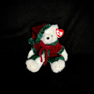 May include: A white plush bear wearing a red velvet outfit with green trim and a matching hat. The bear has a red bow and a red heart-shaped tag. A festive holiday-themed plush toy.