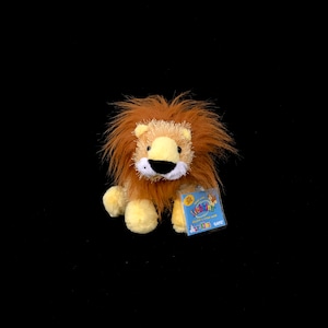Puede incluir: Un león de peluche con una melena marrón y desgreñada, patas amarillas y un hocico blanco. El juguete tiene una etiqueta con las palabras "Webkinz" y "Lil'Kinz". El león está sobre un fondo negro.