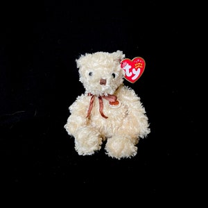 Puede incluir: Un pequeño oso de peluche color crema con una nariz marrón y una cinta marrón. Tiene una etiqueta roja en forma de corazón que dice "Beanie Babies". El oso mide aproximadamente 15 cm de altura. Textura suave y esponjosa.